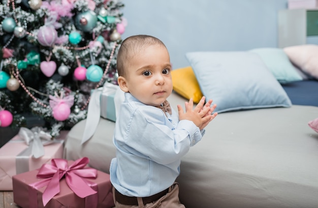 A little boy in a blue shirt stands in a room near the bed against  a Christmas tree