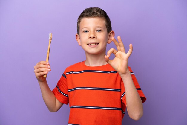 Little boy brushing teeth isolated on purple background showing ok sign with fingers