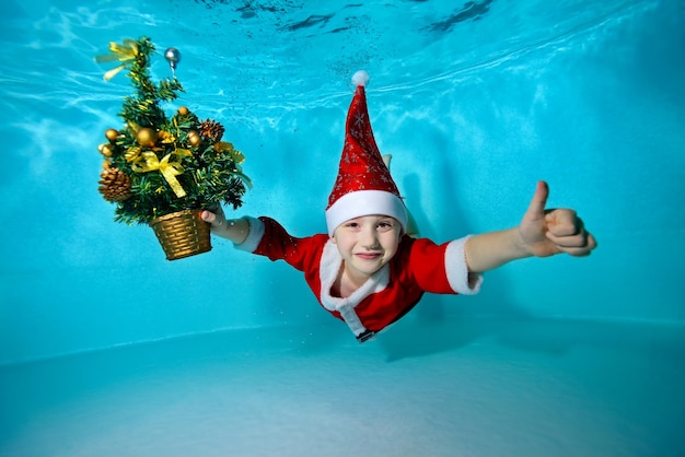 A little boy in a cap Santa Claus swimming and posing underwater with a Christmas tree in hand