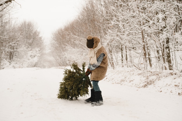 A little boy carries a Christmas tree in an elf costume in a snowy forest. A dwarf in a fur coat is dragging a tree along a snowy road