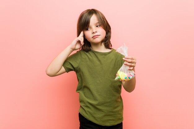 Little boy caucasian holding candies pointing his temple with finger