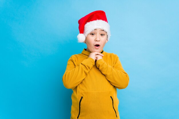 Little boy celebrating christmas day wearing a santa hat isolated praying for luck, amazed and opening mouth looking to front.