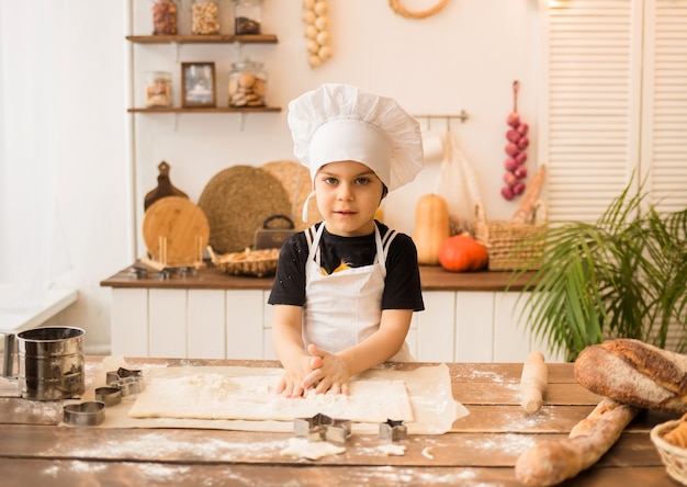 A little boy in a chef's hat and apron prepares dough at a wooden table in the kitchen