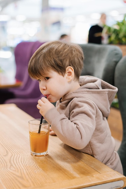 小さな男の子の子供は、カフェのテーブルに座ってストローからジュースを飲みます