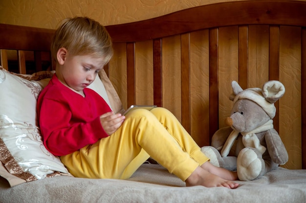 Маленький мальчик в детской комнате на диване смотрит мультфильмы на планшете перед сном