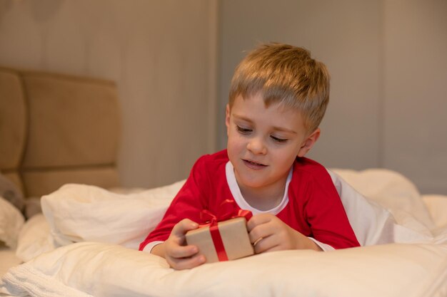 A little boy in Christmas pajamas unpacks a gift while lying in bed Christmas and children concept