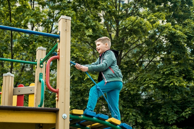 Маленький мальчик лазит по веревке на детской площадке на открытом воздухе. мальчик 6-7 лет на детской площадке лазает по веревке, веселое детство в детском саду и школе, прогулка летом.