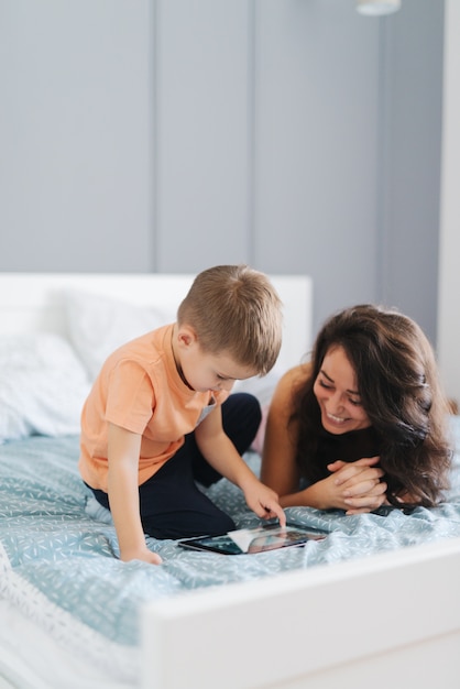 Little boy crouching on the bed and watching cartoons on tablet while his mother lying down and watching cartoons, too. bed time concept. Bedroom interior.