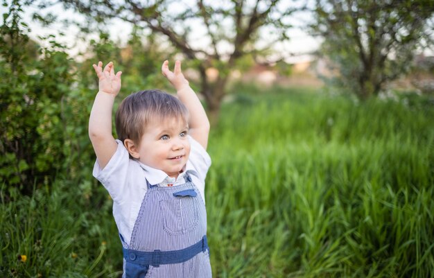 表情豊かな青い目をしたデニムのオーバーオールの男の子。大きな緑の茂みと咲く家の庭を背景に、背の高い緑の芝生でジャンプして浮気します。