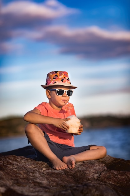 Little boy drinking coconut sitting on the rock near a sea