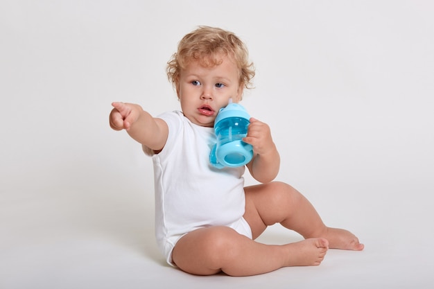 Little boy drinking water while sitting on floor isolated over white space, looking and pointing away, wearing body suit