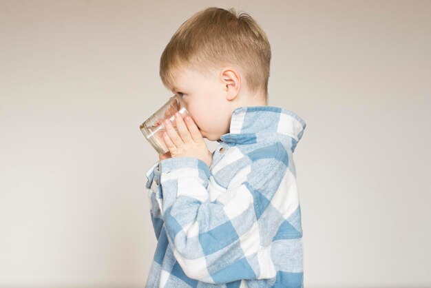 スタジオの灰色の背景のグラスから水を飲む小さな男の子