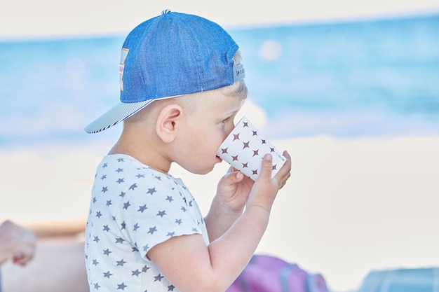 小さな男の子がビーチでプラスチックガラスから水を飲む高品質の写真