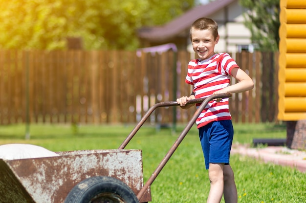 小さな男の子が庭で晴れた日にバッグを持って手押し車を運転します。