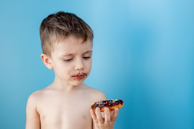 ドーナツチョコレートを食べる少年。かわいい幸せな少年が彼の口の周りにチョコレートを塗った。子供のコンセプト、子供のためのおいしい料理