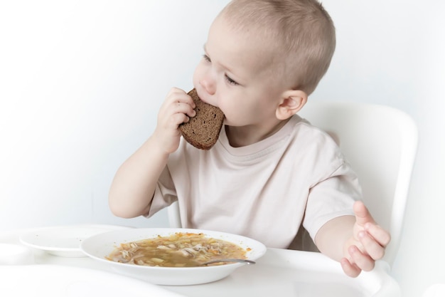 A little boy eats soup with bread on his own in a highchair against the background of a white wall