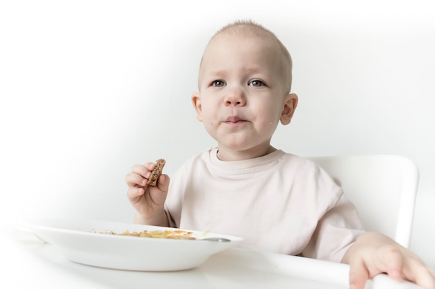 小さな男の子が白い壁を背景にハイチェアで自分でパンと一緒にスープを食べる