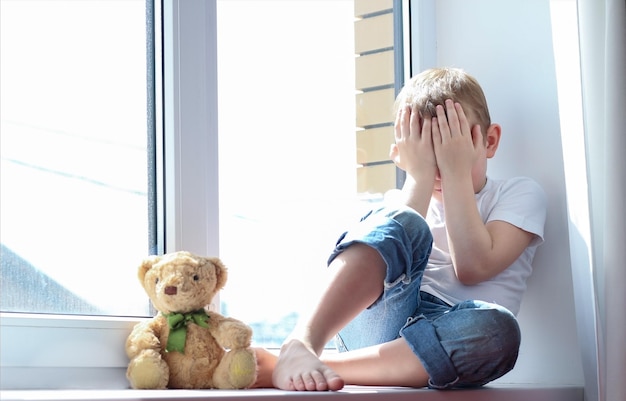 Photo a little boy fairhaired sits on the window and is sad perhaps he misses his father or mother or someone has offended him and he is upset