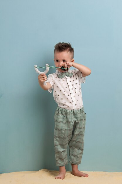 Little boy in fashionable clothing and a slingshot in his hands stands on the sand