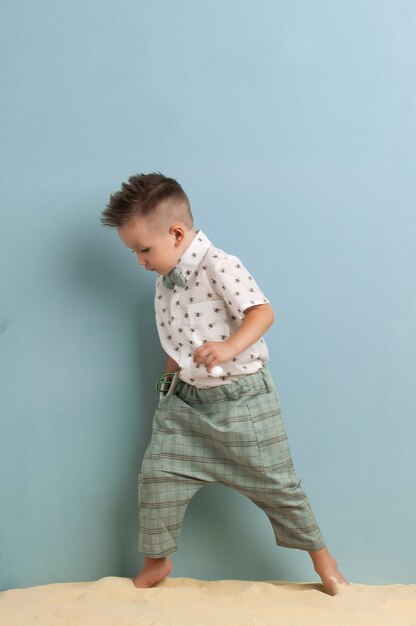 Little boy in fashionable clothing and a slingshot in his hands stands on the sand