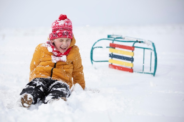 小さな男の子がそりで倒れ、冬の日に雪の子に座る