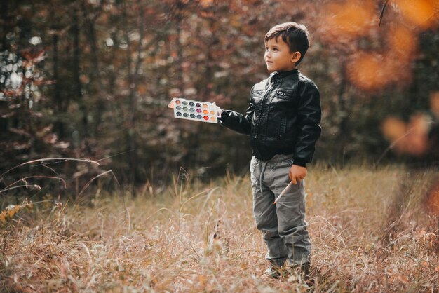 5歳の少年が絵の具を手に森の中に立って、木の葉に絵の具を塗ろうとしています。