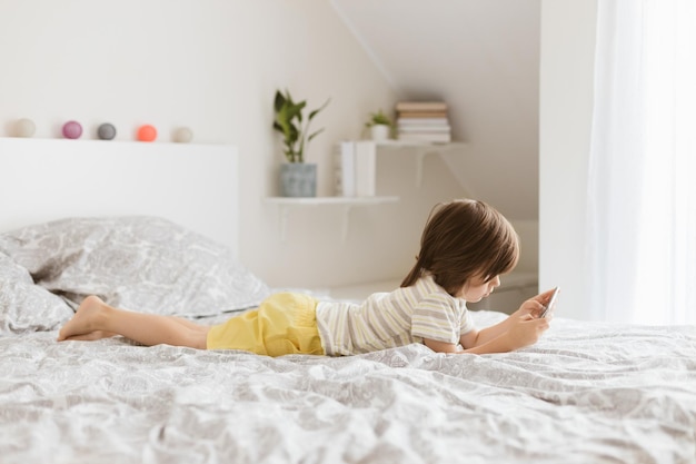 Photo a little boy of four years lies on the bed and plays a mobile phone a smartphone children