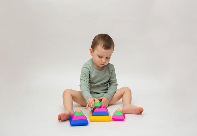 Little boy in a green bodysuit sits and collects a pyramid on a white background