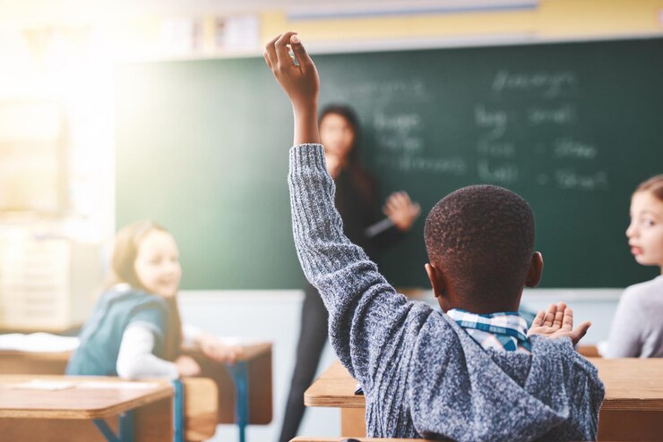 Photo little boy hands raised and classroom with question for teacher answer or learning at elementary school back view kid or young child with answer for interaction engagement or attention in lesson