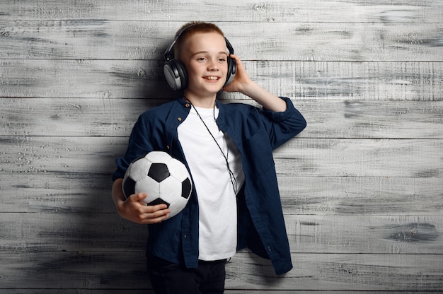 Little boy in headphones holds ball in studio. Children and gadgets, kid isolated on wooden background, child emotion, schoolboy photo session