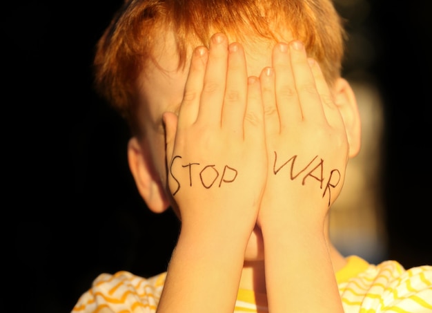 Photo little boy hiding face and words stop war written on his hands outdoors closeup