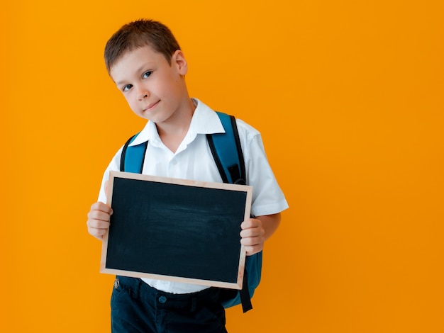 小さな男の子は黄色の背景に学校の黒板を持っています。空白の手でバックパックを持つ小さな小学生の制服。