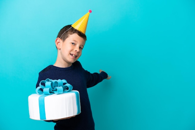 Little boy holding birthday cake isolated on blue background extending hands to the side for inviting to come