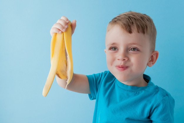 Маленький мальчик держит и ест банан на голубой стене, еде, диете и концепции здорового питания.