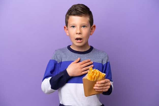 Little boy holding fried chips isolated on purple background surprised and shocked while looking right