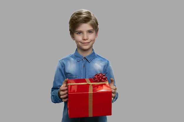 Маленький мальчик держит большую подарочную коробку. Стильный маленький мальчик, держащий настоящую коробку на сером фоне. Получите концепцию праздничного бонуса.