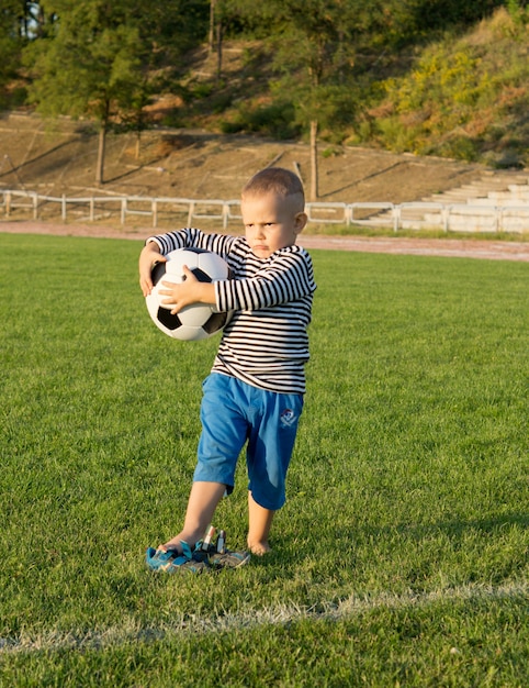 Little boy holding a soccer ball on a grassy sportsfield looking into evening sunlight