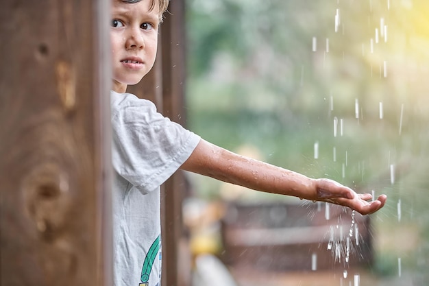 Little boy holds hand under falling down rain drops standing on country house terrace