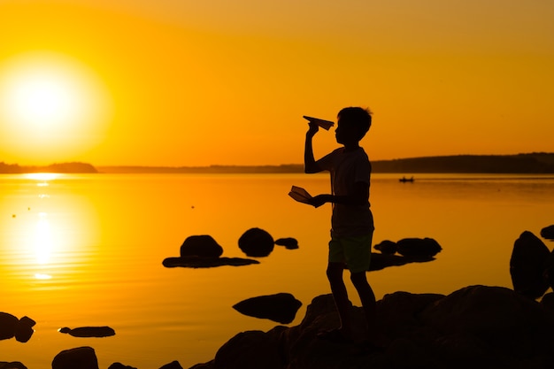 Little boy holds a paper plane in hand at sunset. a child\
raised his hand up to sky and plays with origami in the evening at\
lake. silhouette