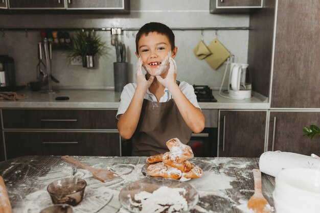 A little boy at home in the kitchen sculpts dough on the table and laughs. The kid is happy that he is soiled in flour.