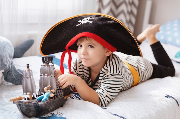Фото Маленький мальчик в пиратском карнавальном костюме, курсирующем дома с игрушкой пиратский корабль. милый ребенок мальчик весело на хэллоуин или день рождения.
