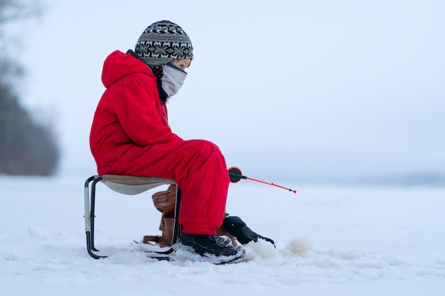 写真 冬の釣りに赤いオーバーオールの小さな男の子。折りたたみ椅子に座っています。釣り竿の手に。