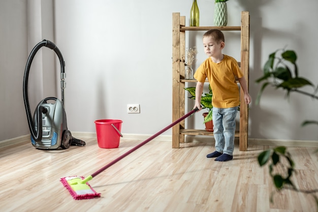 小さな男の子がモップを使って部屋の床を掃除しています。独立の概念、両親への援助、子供の家事