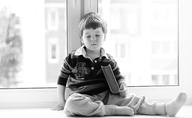 어린 소년이 책을 읽고 있습니다. 아이는 창가에 앉아 수업을 준비합니다. 그의 손에 책을 가진 소년이 창턱에 앉아 있다.