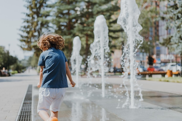 Little boy is running in the street and playing with the water jets of a fountain spouting from the ground. child in blue T-shirt and white shorts is fooling around outdoors. lifestyle. space for text