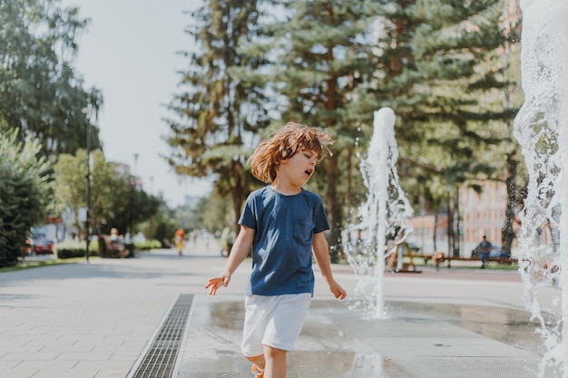 Little boy is running in the street and playing with the water jets of a fountain spouting from the ground. child in blue T-shirt and white shorts is fooling around outdoors. lifestyle. space for text