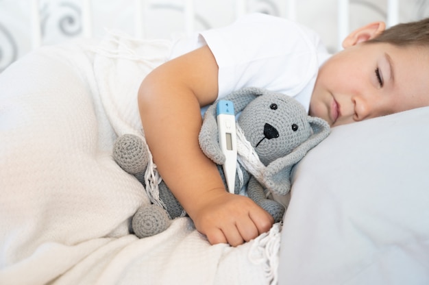 The little boy is sick in bed with a thermometer and high temperature. Sleeping boy lay on the bed.