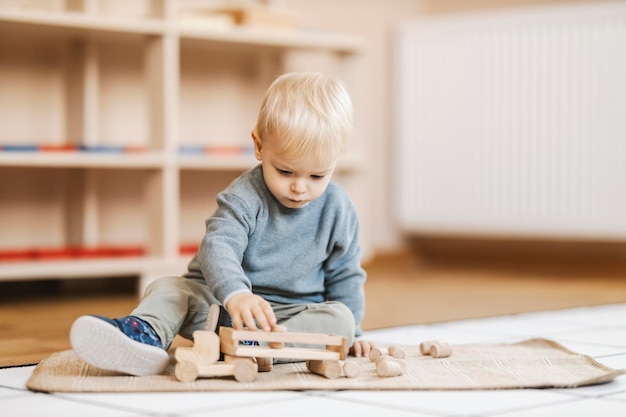 小さな男の子が幼稚園の床に座って、木製のトラックで遊んでいます。