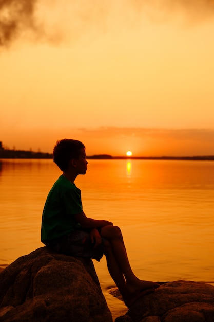 小さな男の子が岩の上に座っています。美しい夏の海の夕日。