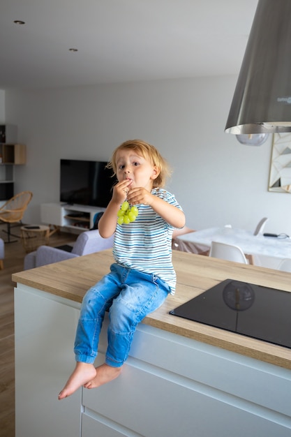 Little boy is sitting on a table in the kitchen and eating grapes. Child is tasting healthy food.